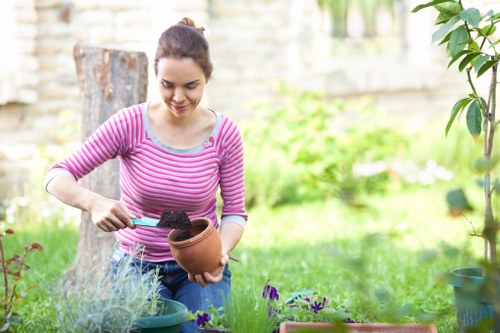 A young woman in a pink and white striped long-sleeve shirt is tending to plants in a backyard garden, holding a terracotta pot filled with soil while using a small garden trowel. The garden features a lush, green lawn and a variety of potted plants, including purple flowering plants and leafy shrubs. In the background, there is a tree trunk, a stone or brick wall, and additional garden greenery, suggesting a well-maintained outdoor space suitable for gardening and landscaping services. The scene is lit with natural daylight, indicating a clear day, and shows an environment typical for outdoor gardening projects in Richmond, where professional gardening services such as hedge trimming and lawn care may be required to maintain the garden's appearance and health.