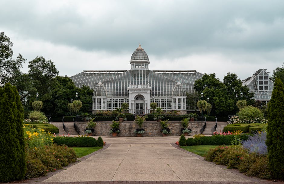 A large Victorian-style glass greenhouse with a metal frame, situated within a lush garden setting. The greenhouse features multiple pitched roofs, some with small decorative cupolas, and is partially
